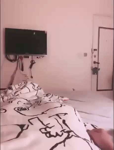 Комната: кровать с рисунчатой простынёй, телевизор на стене и дверь в углу.